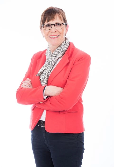 En kvinna med röd kavaj och glasögon som ler stort och har armarna i kors framför sig. 